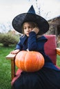 Scary little girl in witch costume, hat with big pumpkin celebrating halloween holiday. Sitting on armchair in coat with pumpkin. Royalty Free Stock Photo