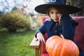 Scary little girl in witch costume, hat with big pumpkin celebrating halloween holiday. Sitting on armchair in coat with pumpkin. Royalty Free Stock Photo