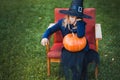 Scary little girl in witch costume, hat with big pumpkin celebrating halloween holiday. Sitting on armchair in coat with pumpkin. Royalty Free Stock Photo