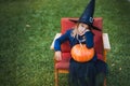 Scary little girl in witch costume, hat with big pumpkin celebrating halloween holiday. Sitting on armchair in coat with pumpkin. Royalty Free Stock Photo