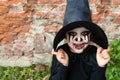 Scary little girl in a witch costume smiles against a brick wall. Happy Halloween