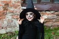 Scary little girl in a witch costume smiles against a brick wall. Happy Halloween