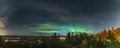 Scenic Aurora Borealis panorama photo across the whole horizon, above autumn birch and pine tree forest, city lights in Swedish