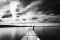 Scenic grayscale view of a wooden dock over a calm lake under a wispy sky