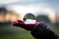 A scenic landscape image reflected inside of a lens ball resting on the red glove of a person. The landscape is a meadow during