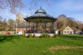 A scenic view of a bandstand in a park with a church and trees in the background under a majestic blue sky