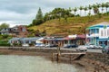 Scenic view of cars parked on the waterfront road in front of shops near a green hill in Mangonui