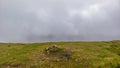 A scenic view of a grassy mountain summit with a stony cairn under a misty cloudy sky