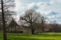 Scenic view of a leafless tree in a lush green field with a traditional thatched-roof cottage in the background under a cloudy sky Royalty Free Stock Photo