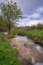 Scenic view of mossy river flowing downstream among grassland with trees on cloudy sky