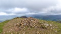 A scenic view of a rocky mountain summit with a stony cairn and mountain range and valley in the background under a stormy grey