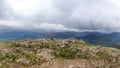 A scenic view of a rocky mountain summit with a stony cairn and mountain range and valley in the background under a stormy grey