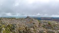 A scenic view of a rocky mountain summit with a stony cairn and mountain range in the background under a stormy grey cloudy sky