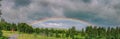 Scenic wide summer rainbow panorama over small mountain village in Scandinavian Mountains. Dramatic rain clouds over forest and Royalty Free Stock Photo