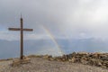 A scenics view of a mountain summit cross with a majestic double rainbow in the background under a stormy and rainy weather