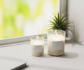 Scented candle with blank label mockup, burning white aromatic candles in glass on white surface with green plant on background, Royalty Free Stock Photo