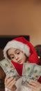 A school-aged girl in a Santa Claus hat, puzzled and surprised, holds cash in dollar bills. Christmas and business Royalty Free Stock Photo