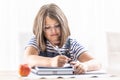 School attending girl wearing glasses writes into an exercise book with a pen having an apple on a table besides her Royalty Free Stock Photo