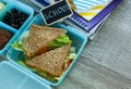 School blue lunch box with homemade sandwich, green apple, cookies, pencils, clock, notebooks on the table.