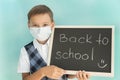 School boy in medical mask holds blackboard with inscription back to school made with piece of chalk.