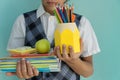 School boy in school uniform holds stack of notebooks, apple, yellow glass with pens and pencils.