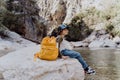 School boy with yellow backpack sits on a riverside rock in the canyon with mountain cliffs in the background. Kid child