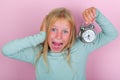 School girl hates waking up early in the morning. Blond girl holding alarm clock covers ear with hand on pink background