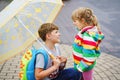 School kid boy picks up the little sister, preschool girl from kindergarten. Two happy children with umbrella playing