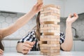 School kids play jenga sitting at kitchen table closeup. Hand of girl put wooden block on top of tower keeping balance.