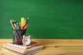 School supplies, pens, pencils, notebooks and an alarm clock on a wooden table against the background of a green school board Royalty Free Stock Photo