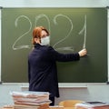 A school teacher in a medical mask writes the number 2021 in chalk on a blackboard. Learning problems during a coronavirus Royalty Free Stock Photo