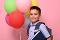 Schoolboy with backpack holding book and multicolored balloons, cute smiling posing to camera over pink background with copy space Royalty Free Stock Photo