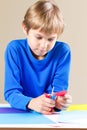 Schoolboy cutting colored paper with scissors at the table