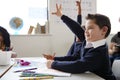 Schoolboy with Down syndrome sitting at a desk raising his hand in a primary school class, close up, side view