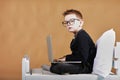 schoolboy in glasses using laptop sitting on bed. Child doing school homework and seriously and tired looking on camera Royalty Free Stock Photo