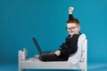 schoolboy in glasses using laptop sitting on bed. happy child doing school homework and joyfully raises his hands up Royalty Free Stock Photo