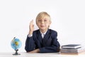 Schoolboy in large glasses sits at table with textbooks with his hand raised. Excellent student. Boy knows the answer