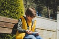 Schoolboy plays game his cell phone sitting on bench in the city park. Boy kid in yellow vest and green hoodie uses