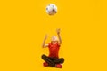 Schoolboy sits cross-legged and tosses soccer ball up. Yellow background. Footballer child with ball