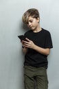 A schoolboy stands with a phone in his hands against a gray wall. A handsome guy in a black t-shirt and green pants. Chats, games