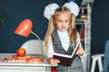 Schoolgirl blondy in school uniform with white bows standing near the table with red lamp and Apple and studying at home.