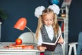 Schoolgirl blondy in school uniform with white bows standing near the table with red lamp and Apple and studying at home.