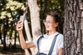 A schoolgirl in glasses takes a selfie on a smartphone near a tree in the park