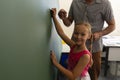 Schoolgirl with math teacher looking at camera and writing on chalk board in classroom Royalty Free Stock Photo