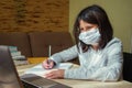 A schoolgirl with a medical mask on her face watches an online lesson and takes notes. The concept of home schooling during