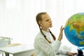 A schoolgirl with pigtails and a white shirt sitting at her desk is looking at a globe in a school classroom. The