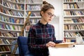 Schoolgirl in a plaid shirt leafing through a book while sitting in a library room. Young woman came to the library to prepare for