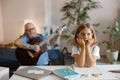 Schoolgirl sits at table while daddy bothers her playing guitar on sofa in living room Royalty Free Stock Photo