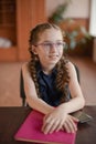 Schoolgirl sitting at the table with textbooks in the classroom. Girl in a blue dress with pigtails