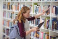 Schoolgirl using digital tablet while selecting book in library Royalty Free Stock Photo